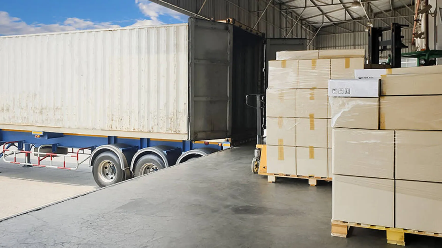 Stacked pallets of boxes on a warehouse loading dock beside an open trailer, ready for shipment